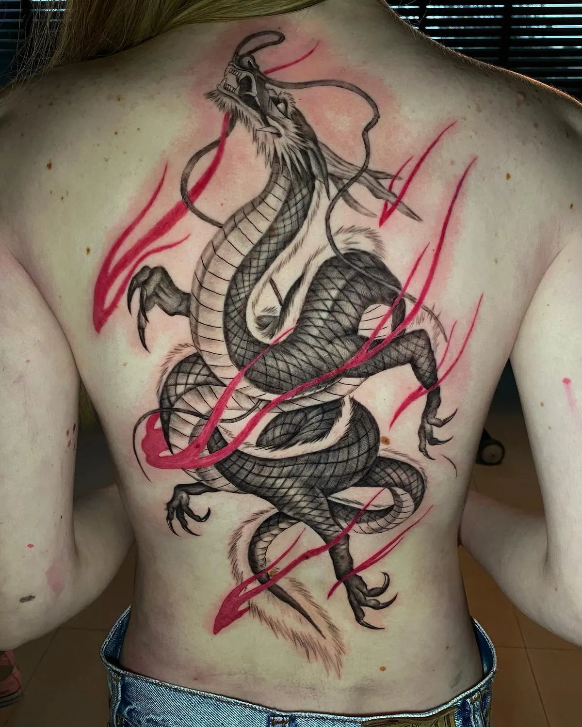 Dragão oriental tatuado nas costas inteiras de uma mulher de cabelo loiro, em preto e cinza, com rastros vermelhos ao redor