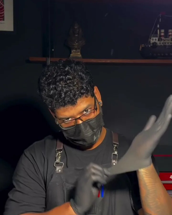Lucas de máscara e avental calçando luvas pretas, com a estante do estúdio ao fundo