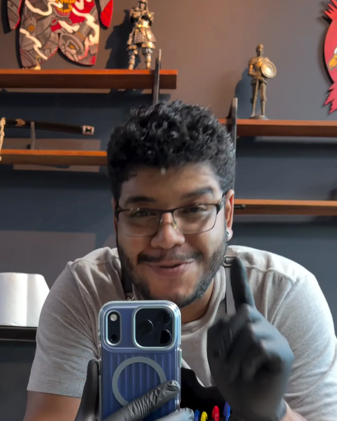 Lucas Lopes no estúdio, de óculos, luvas pretas e avental, sorrindo e apontando para cima, com prateleiras de miniaturas atrás